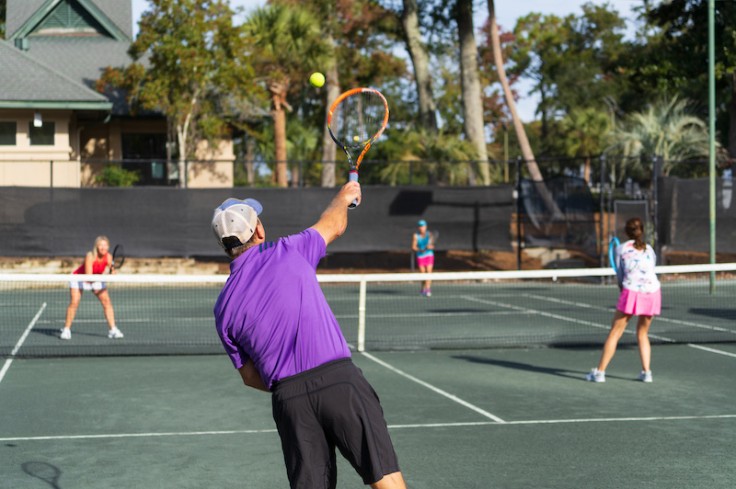 Male tennis player mid serve on the tennis court with other players in the background at Palmetto Dunes Tennis Center