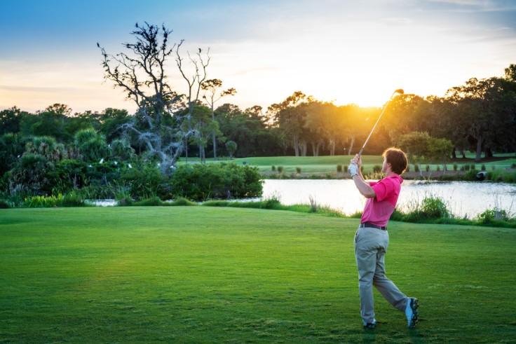 Male in a pink shirt post swing on the George Fazio golf course with a sun flare and water behind him