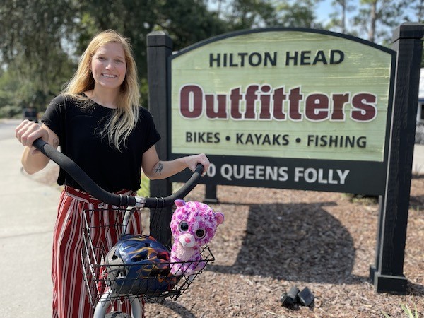 Palmetto Dunes employee riding a bike in front of the Hilton Head Outfitters sign with Purple in the basket