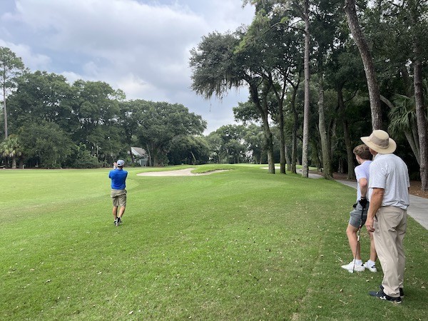 male golfer post swing observing his drive with two male golfers on the sidelines watching at Palmetto Dunes Golf Academy
