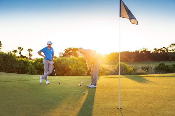 two male golfers putting into the hole with a yellow flag and sun glare behind them