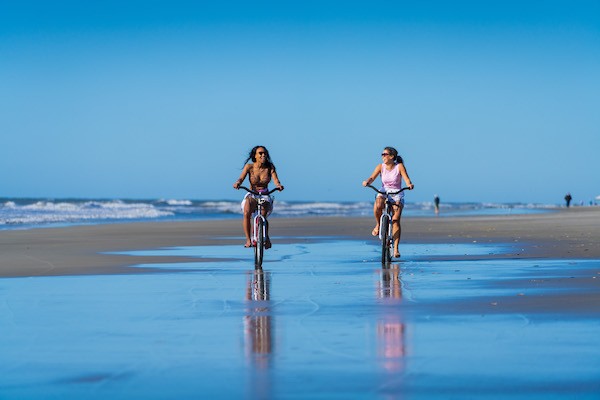 two women riding bikes on the beach with water and sand surrounding them