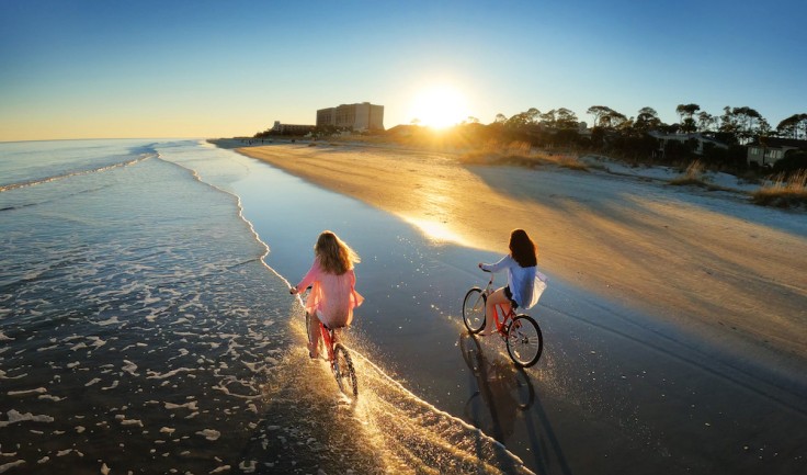 two women on bikes on the beach with the sun setting in the background