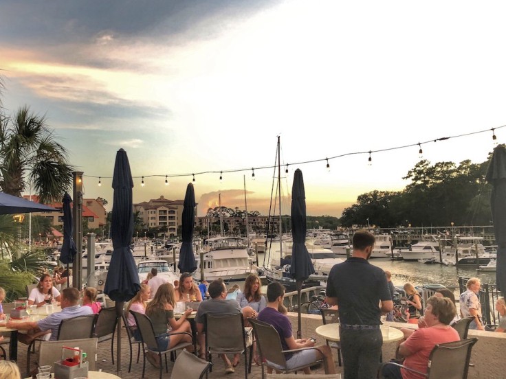 people dining outdoors overlooking marina and boats with the sun setting in the background