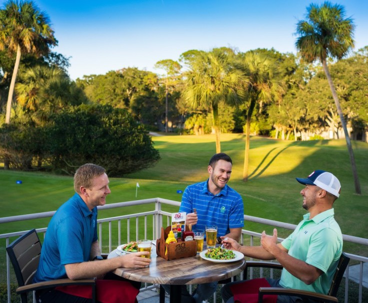 3 men in golf apparel laughing over drinks and food overlooking golf course