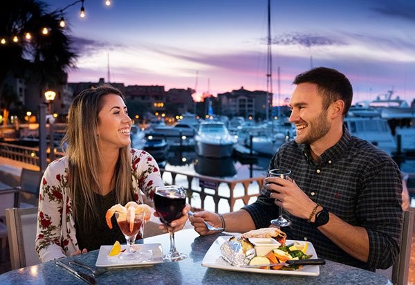 Couple laughing with wine at dinner with marina and sunset in background