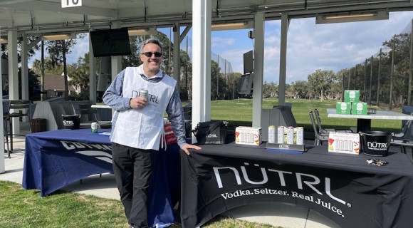 man wearing a Nutrl vest leaning against a Nutrl seltzer promo table with Toptracer Driving Range in the background