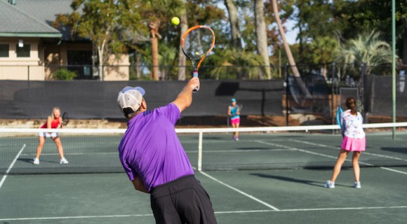 Male tennis player mid serve on the tennis court with other players in the background at Palmetto Dunes Tennis Center