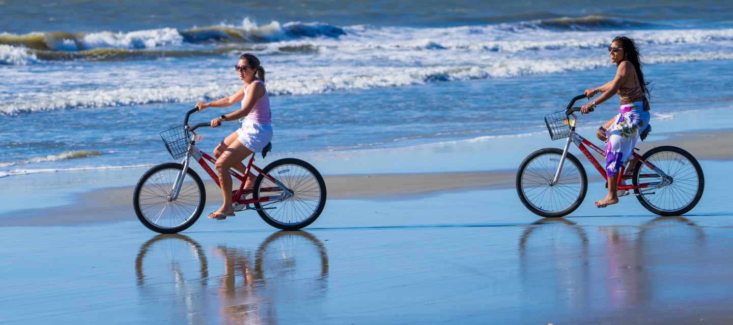 two women riding bikes on the beach with waves in the background