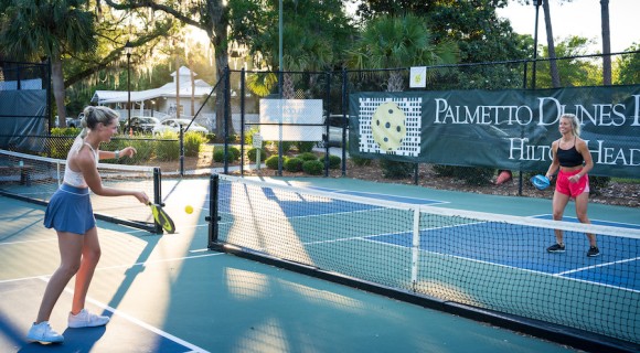 two young women actively playing pickleball at Palmetto Dunes Pickleball court with the sun shining through the trees