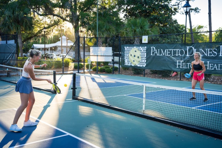 two young women actively playing pickleball at Palmetto Dunes Pickleball court with the sun shining through the trees