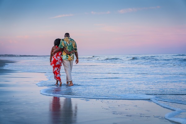 couple in floral clothing embracing each other as they walk along the water with the sunset on the beach