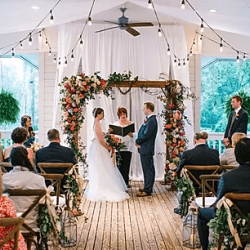 wedding ceremony with bride and groom under a flowered arch with guests seated in wooden chairs and lights above