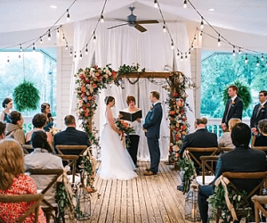 wedding ceremony with bride and groom under a flowered arch with guests seated in wooden chairs and lights above