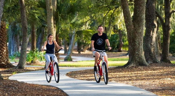 A man and a woman biking on paved path with trees surrounding them.