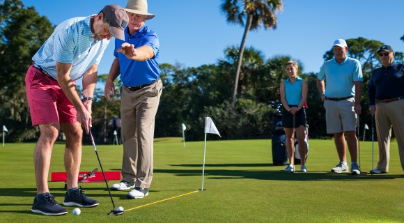 Golf instructor with golfer on putting green with three golfers watching.