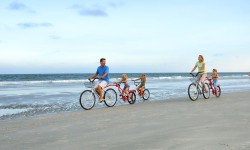 Family of four riding bikes on beach near ocean on clear day.